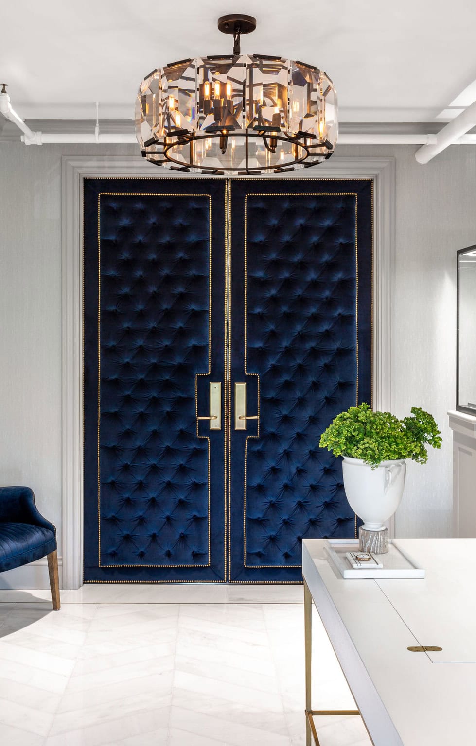 Double doors upholstered in blue velvet with gold trim and rectangular handles, beneath a modern chandelier. A white table with a potted plant sits in the foreground.