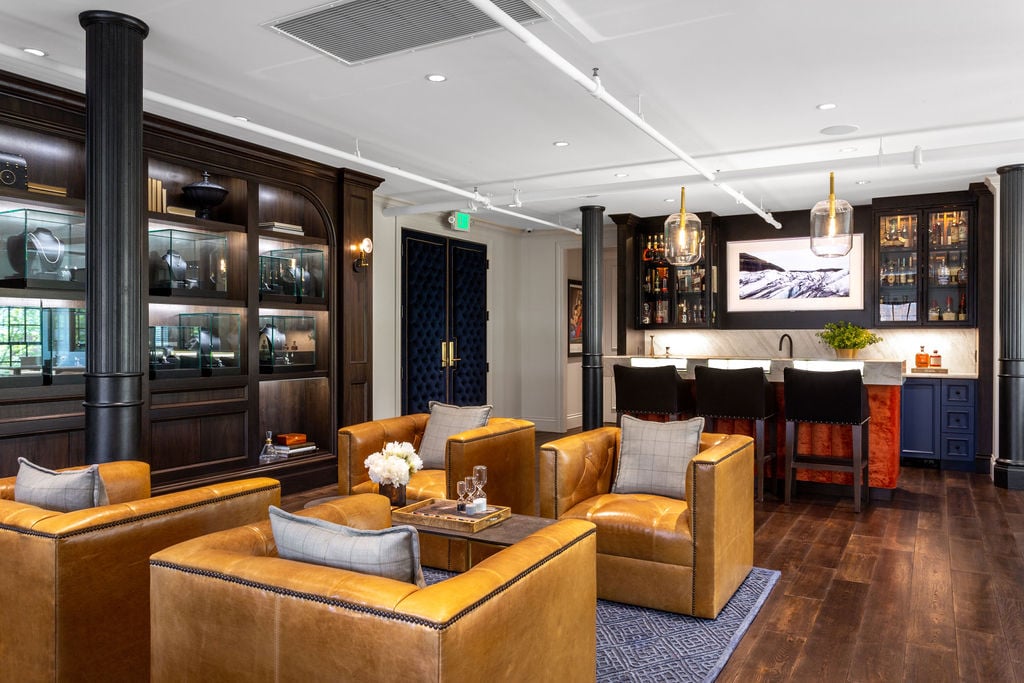 Modern lounge with brown leather armchairs, dark wood floors, a built-in bar, display shelves, and framed art above the bar area.