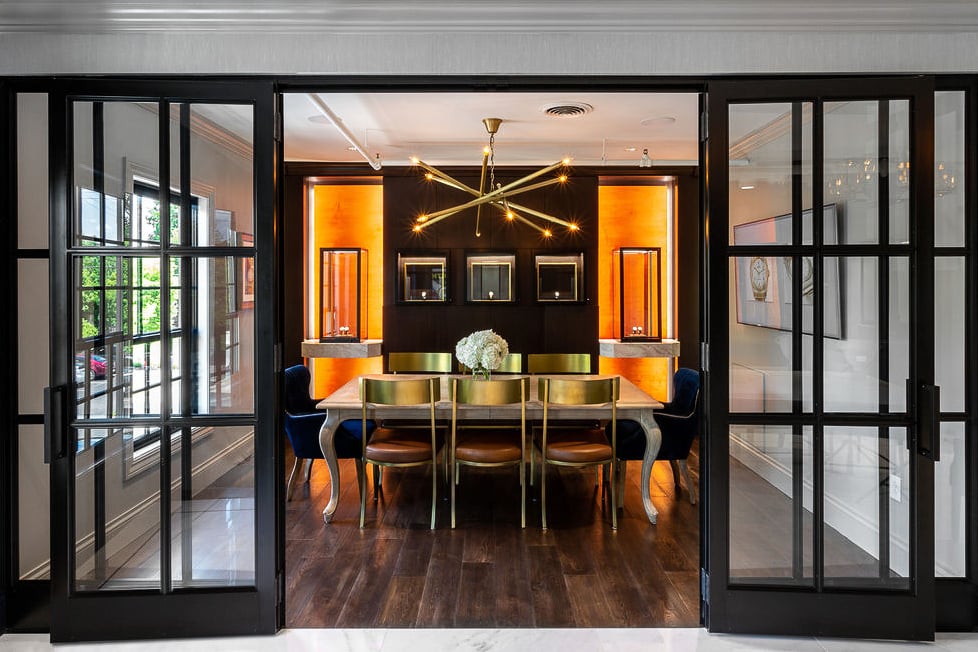 Modern dining room with a long wooden table, gold chairs, central floral arrangement, contemporary chandelier, and glass sliding doors. Warm lighting highlights wall shelves in the background.