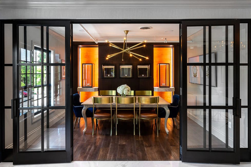 Modern dining room with a long wooden table, gold chairs, a central floral arrangement, geometric chandelier, and glass sliding doors. Warm backlighting highlights wall displays.