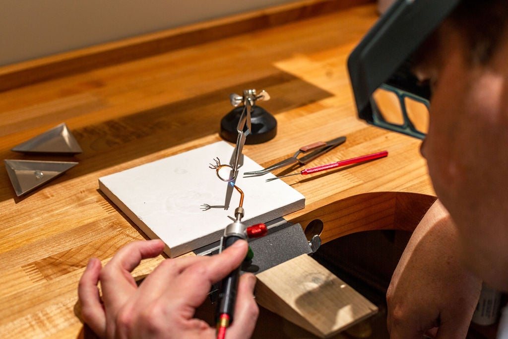 Person using a magnifier and tools at a workbench to hold and work on a small metal sculpture, with tweezers and other tools on the table.