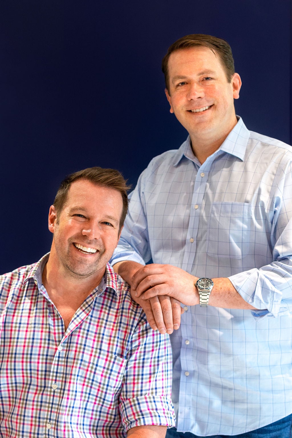 Two men pose together in front of a dark blue background, one seated and smiling, the other standing with his hand resting on the seated man's shoulder.