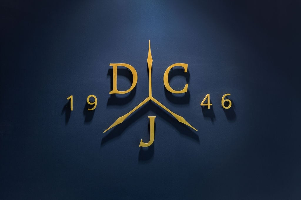 Gold letters and numbers "D", "J", "C", "1946" arranged around three stylized pen nibs on a dark blue background.