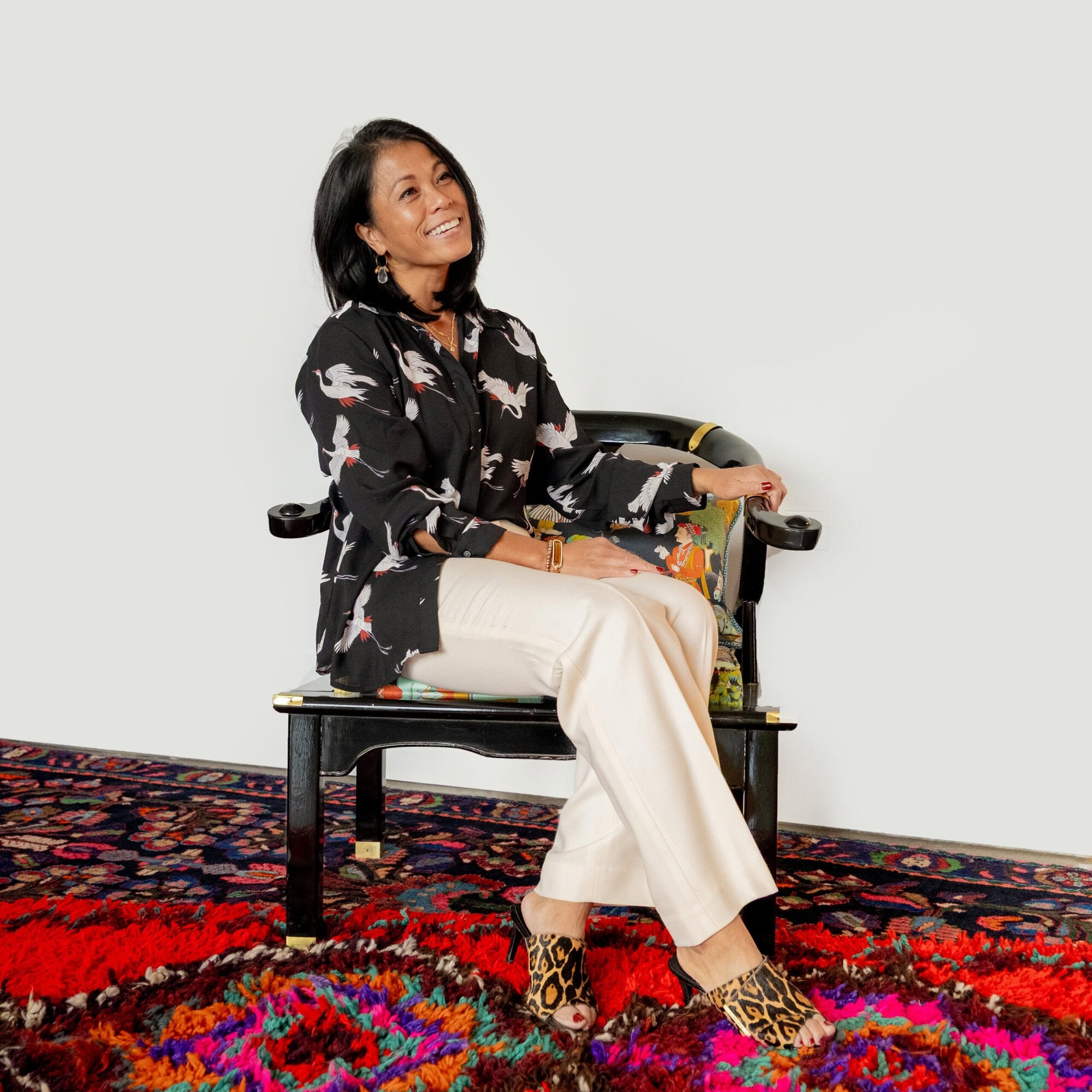 A smiling woman wearing a crane-print shirt and white trousers sits in a black chair on a colorful patterned rug.