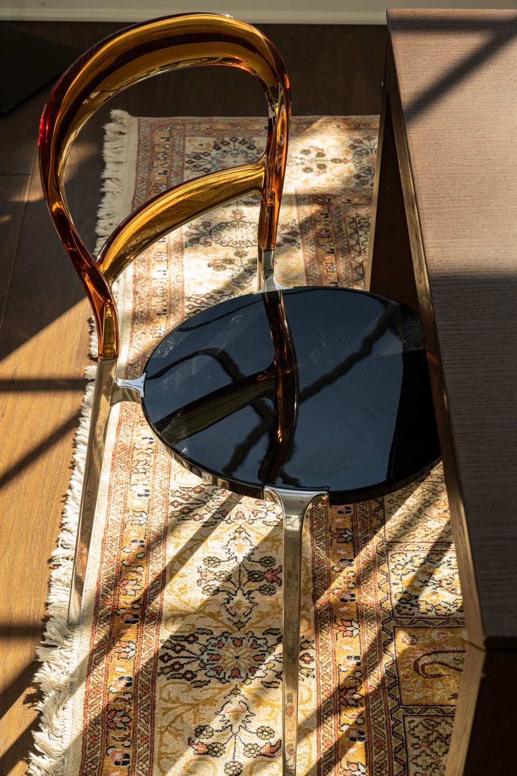 A modern chair with an amber back and black seat sits on a patterned rug next to a dark wooden table, illuminated by sunlight.