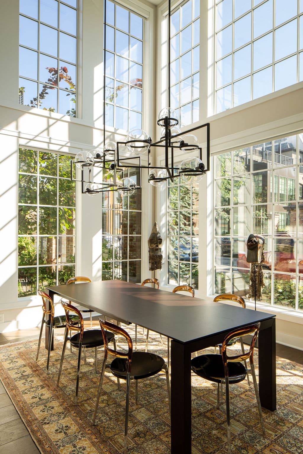 Modern dining room with large floor-to-ceiling windows, a black rectangular table, eight chairs, a geometric chandelier, and a patterned rug, filled with natural light.