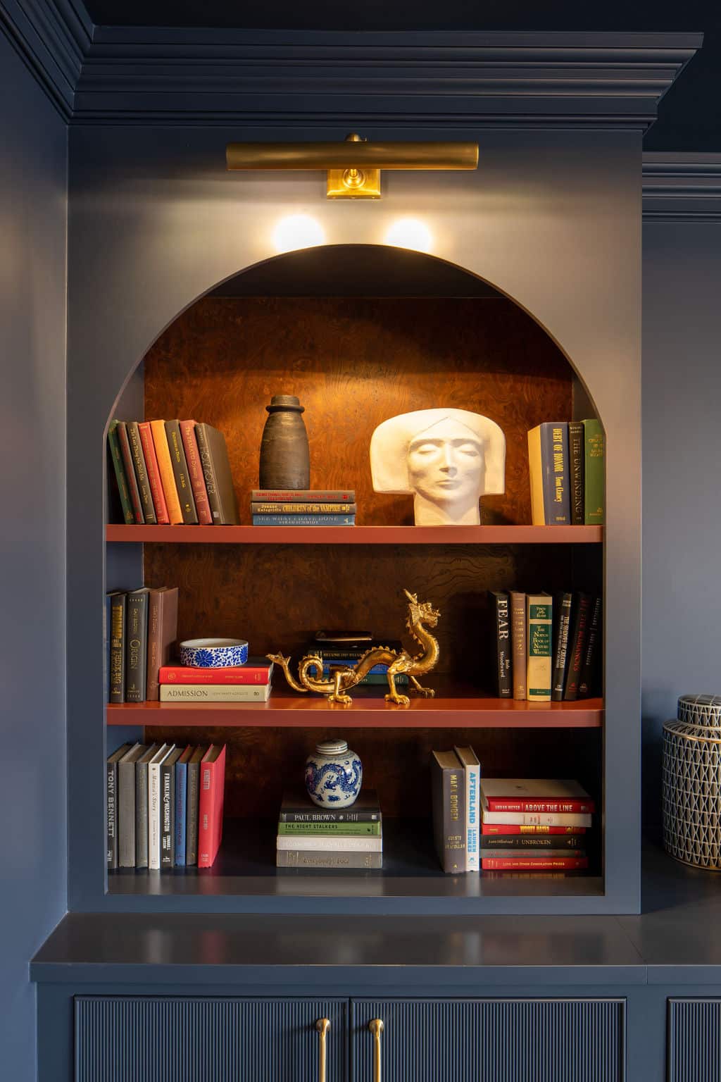 A built-in arched bookshelf by Crimson Design Group displays books, a ceramic vase, a white sculpted head, a blue and white jar, a gold dragon figurine, and a small blue dish, all illuminated by an overhead light.