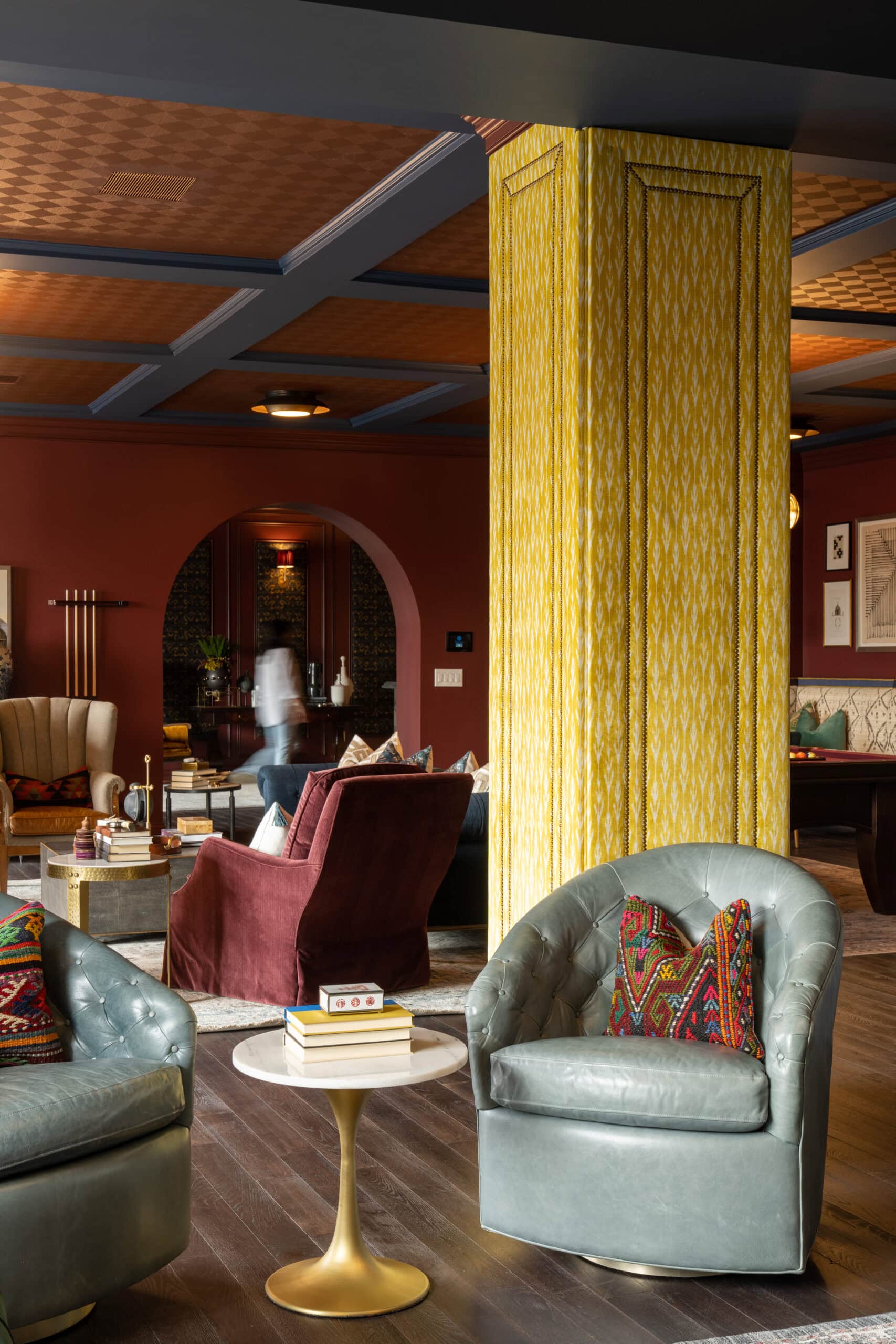A stylish lounge with velvet armchairs, patterned pillows, a small round side table with books, and a yellow pillar, featuring warm lighting and eclectic decor.
