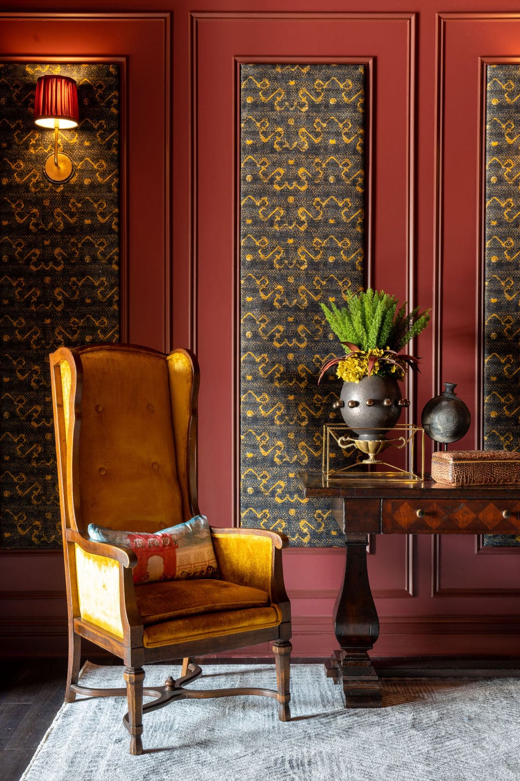 A yellow velvet armchair sits beside a wooden table with a potted plant and decorative items, against a red paneled wall with patterned wallpaper.