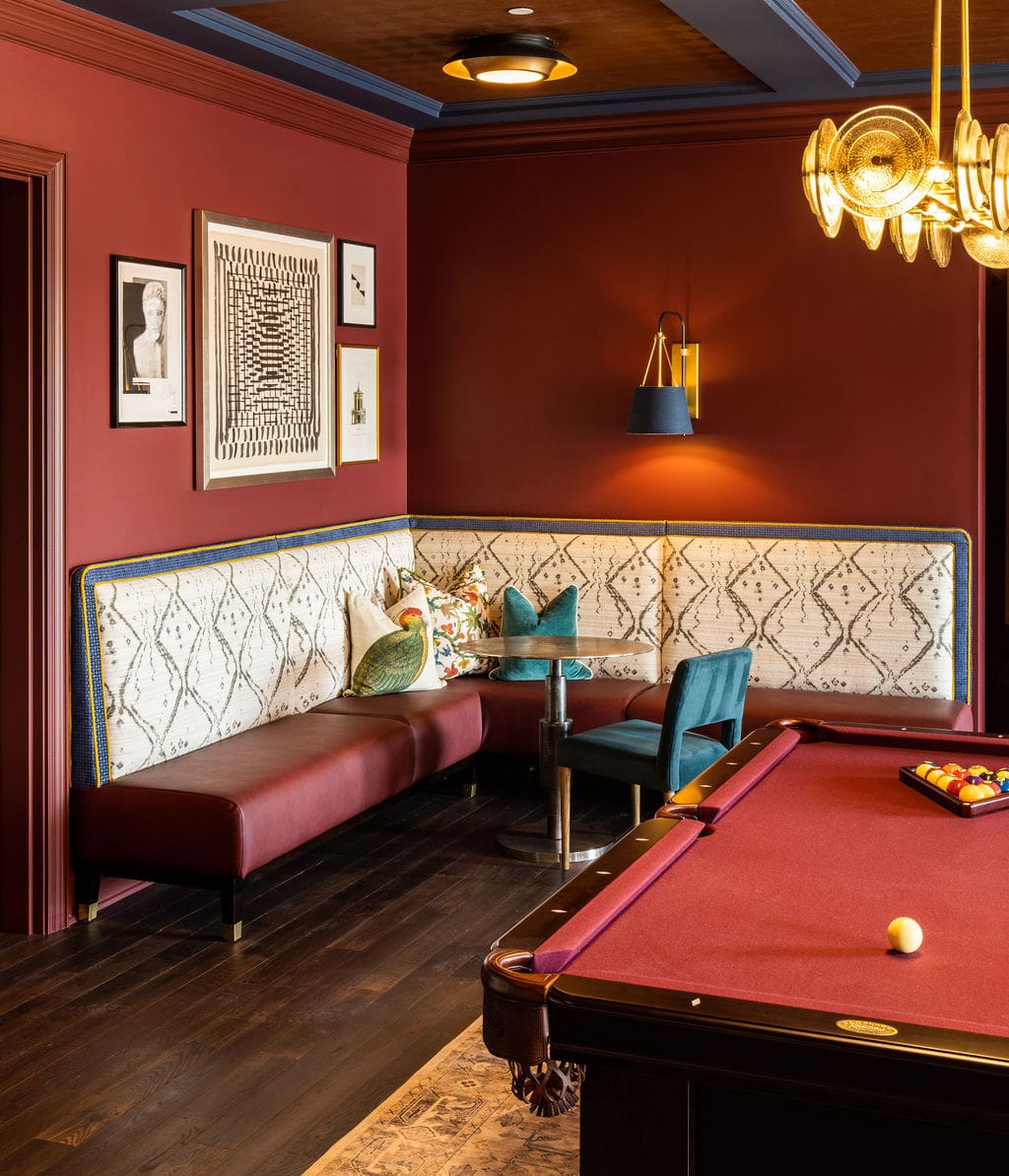 A corner seating area with patterned upholstery, a small round table, teal chair, and wall art, next to a red pool table in a room with dark wood floors and burgundy walls.