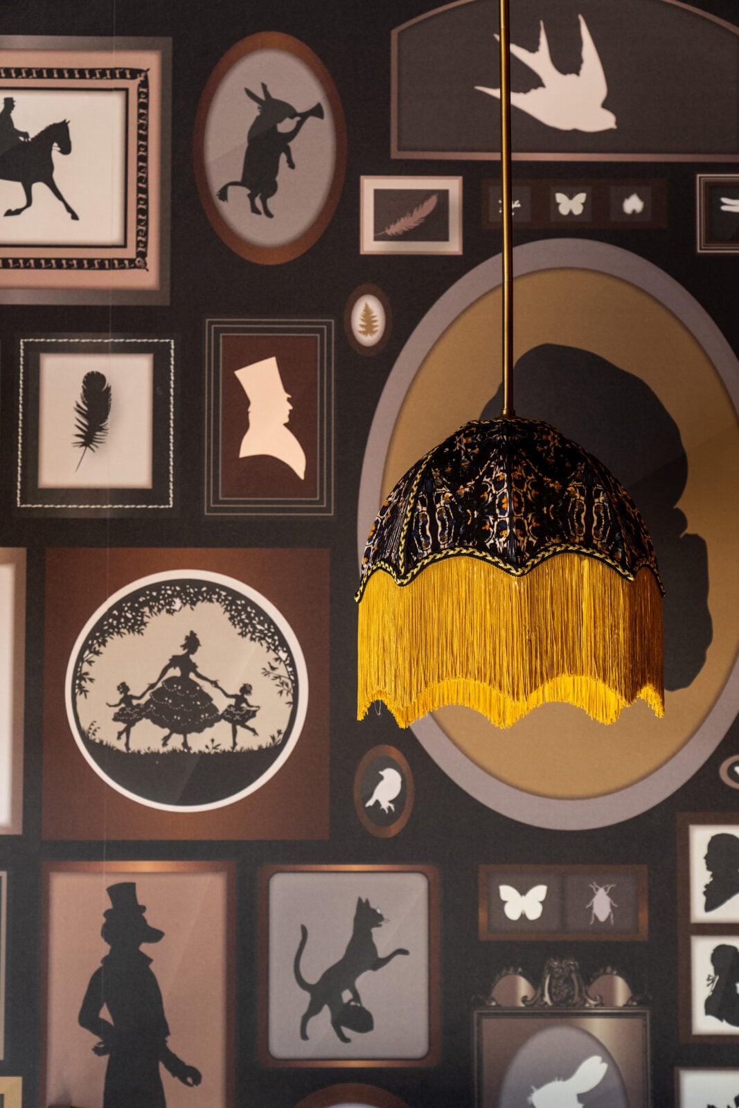 Pendant lamp with yellow fringe hangs in front of a wall decorated with framed silhouette art and animal illustrations in various shapes and sizes.