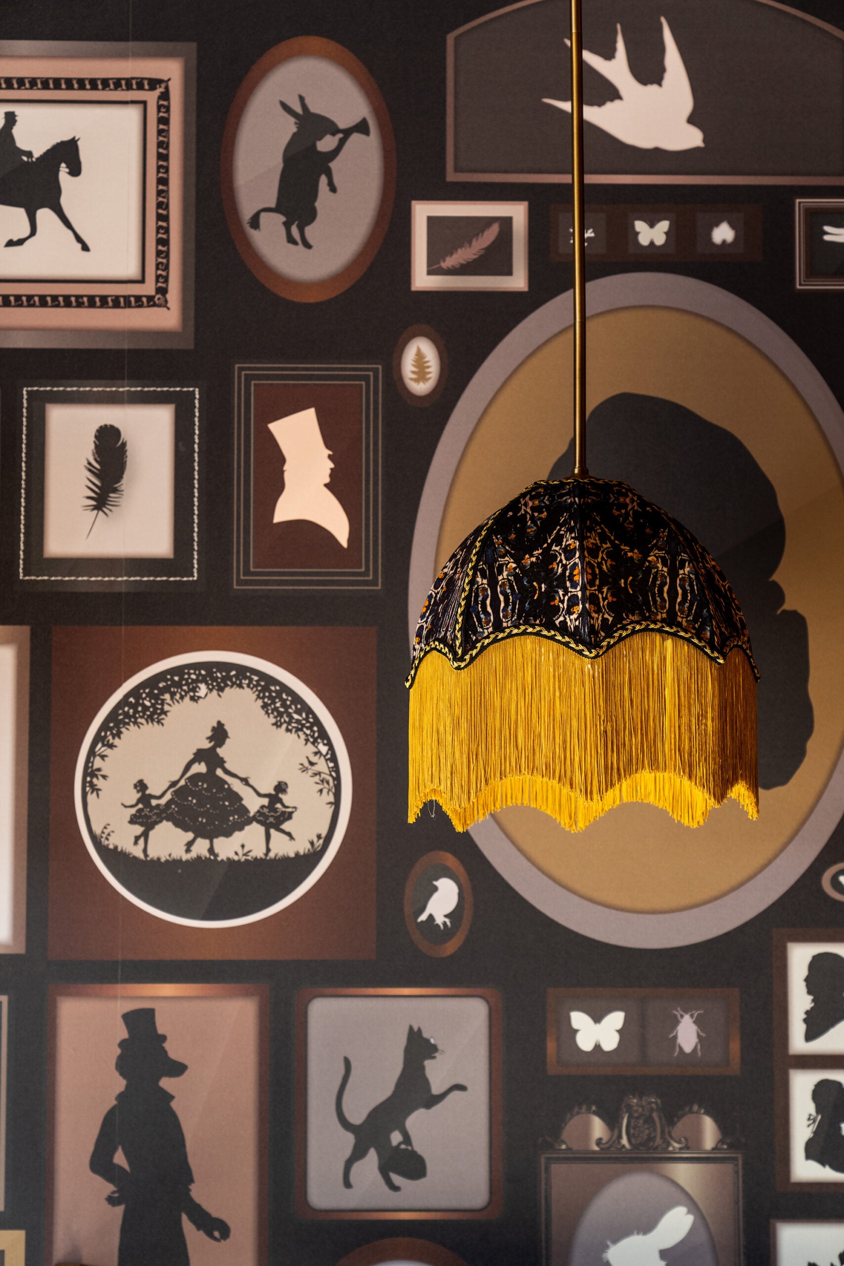 Pendant lamp with black and gold fringe hangs in front of a wall covered in silhouette artwork, including animals, people, insects, and decorative frames.