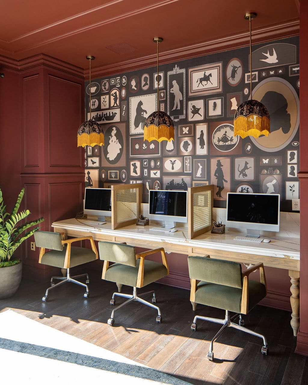 A row of four computers with privacy dividers on a wooden desk, green chairs, and three pendant lights, set against a wall with framed art silhouettes.