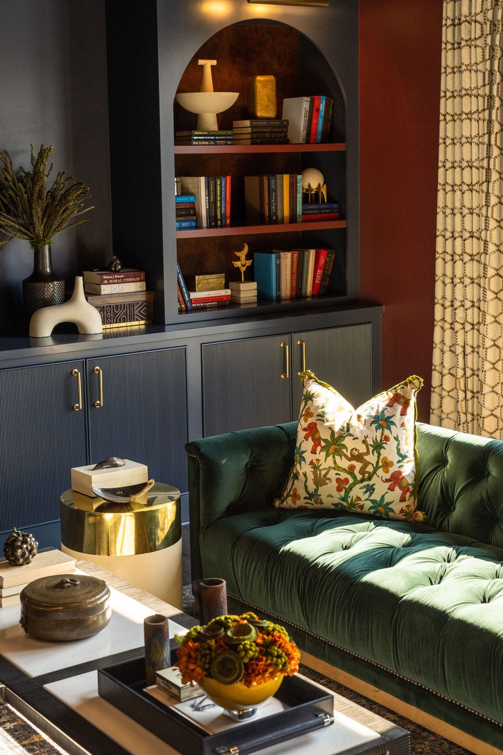 A living room with a green tufted sofa, floral pillow, gold side table, white coffee table, and built-in navy shelves filled with books, decor, and ceramics.