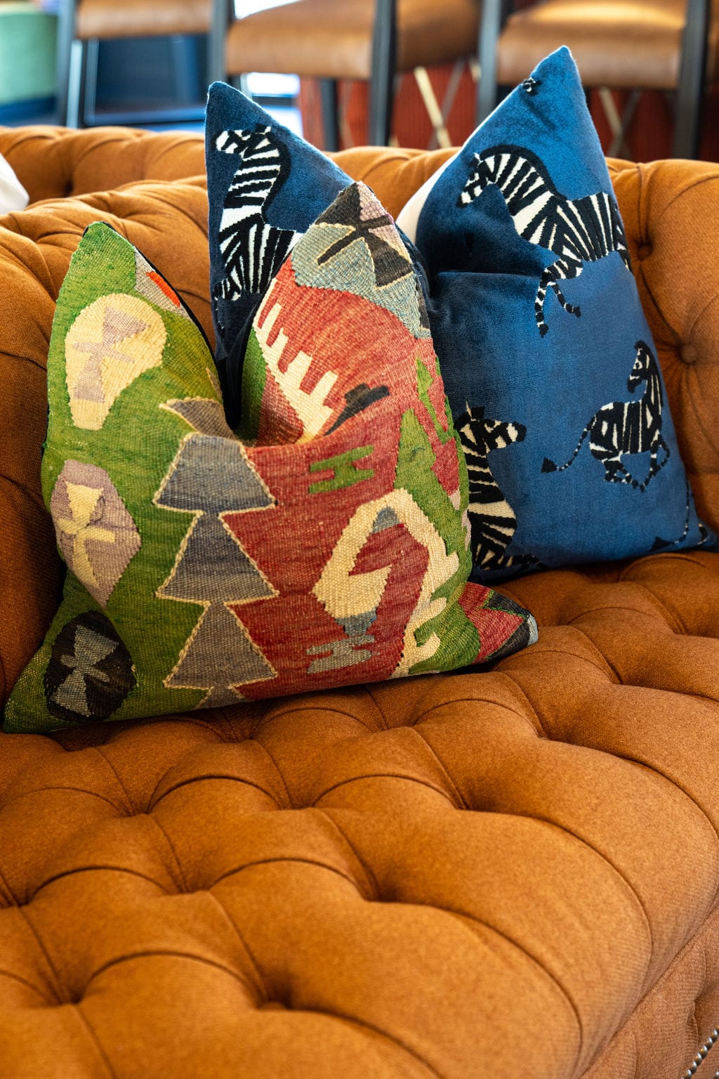 Two patterned throw pillows, one with a geometric design and the other with zebras, by Crimson Design Group, are placed on a tufted brown sofa.