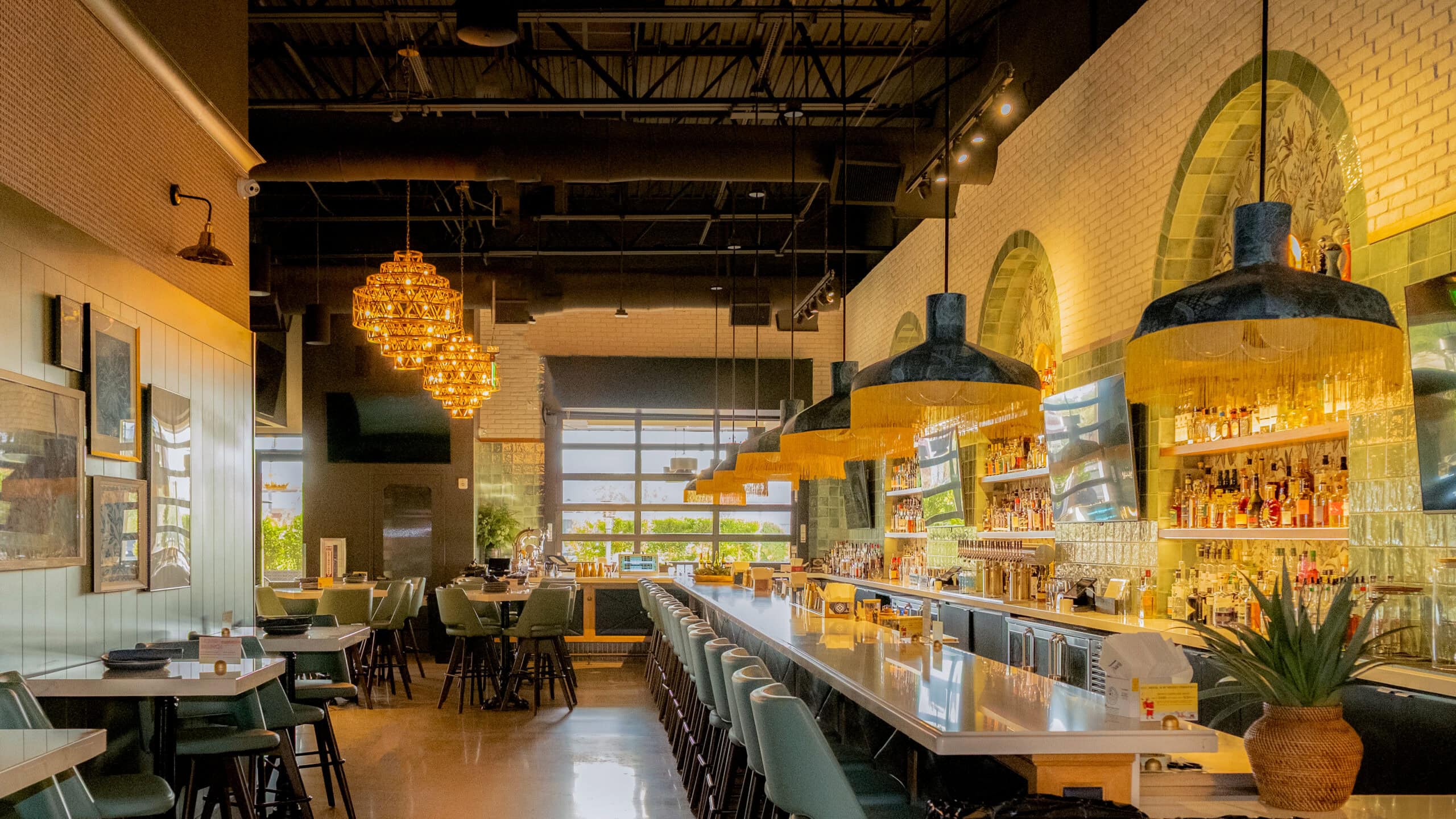 Modern restaurant interior with a long bar, green chairs, hanging lights, and large windows letting in natural light. Tables are set and shelves behind the bar display bottles.