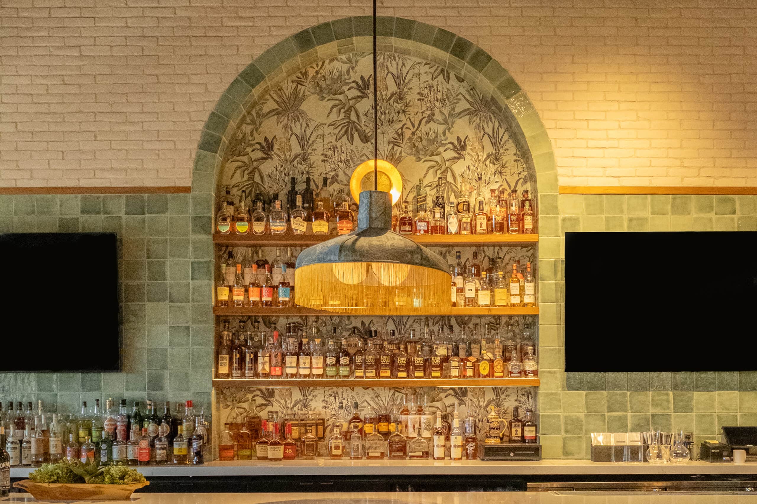 A bar with shelves stocked with various bottles of liquor, a central hanging light fixture, and two flat-screen TVs mounted on a tiled wall.