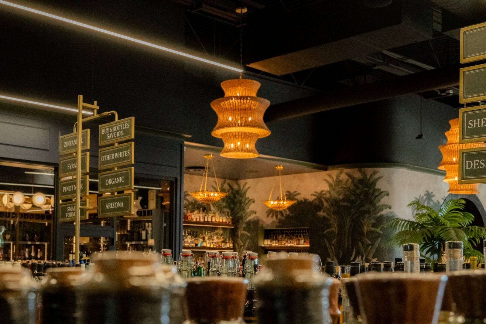 Bar interior with assorted bottles on the counter, hanging chandeliers, decorative signage, and a tropical-themed mural on the back wall.