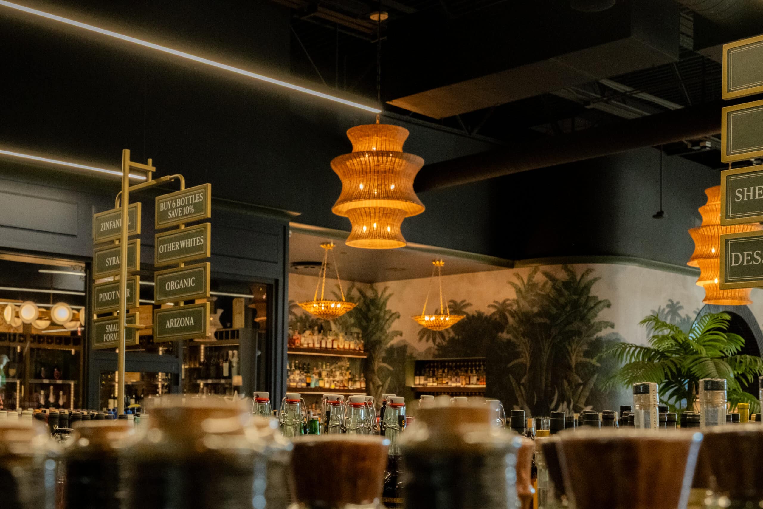 Bar interior with assorted bottles on the counter, hanging chandeliers, decorative signage, and a tropical-themed mural on the back wall.