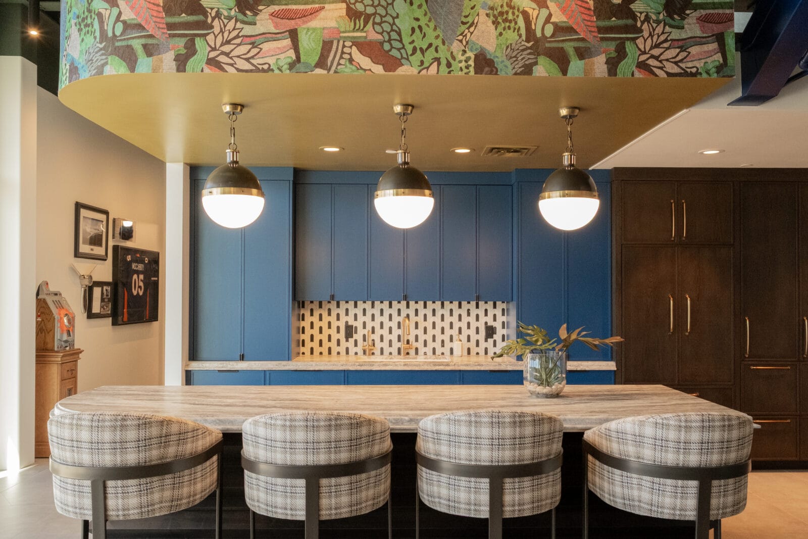 Modern kitchen with a patterned ceiling, blue cabinets, hanging globe lights, a large island with plaid barstools, and a vase with greenery on the countertop.