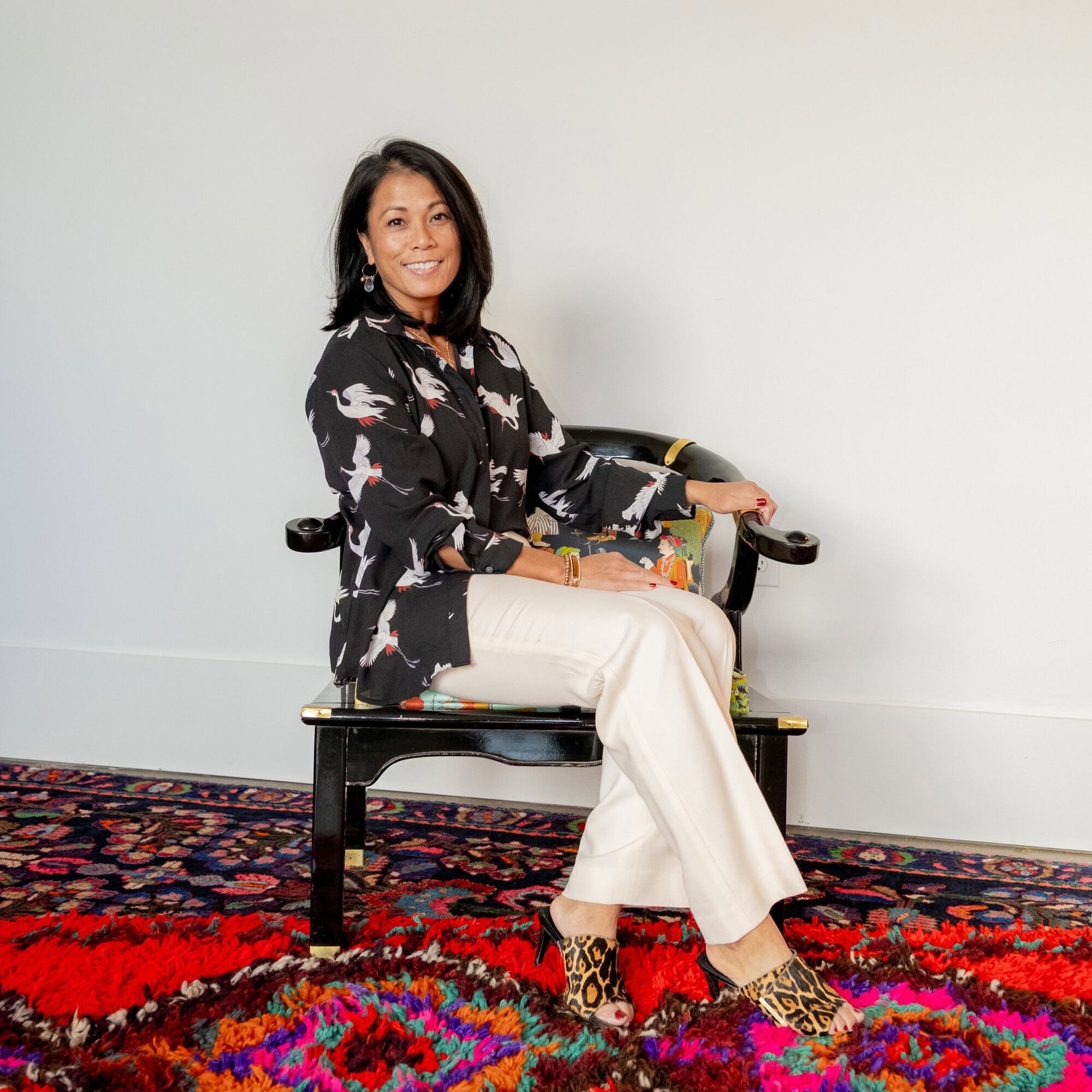 A woman with shoulder-length dark hair sits on a black bench, wearing a patterned shirt, light pants, and leopard-print shoes, on a colorful carpet against a white wall.