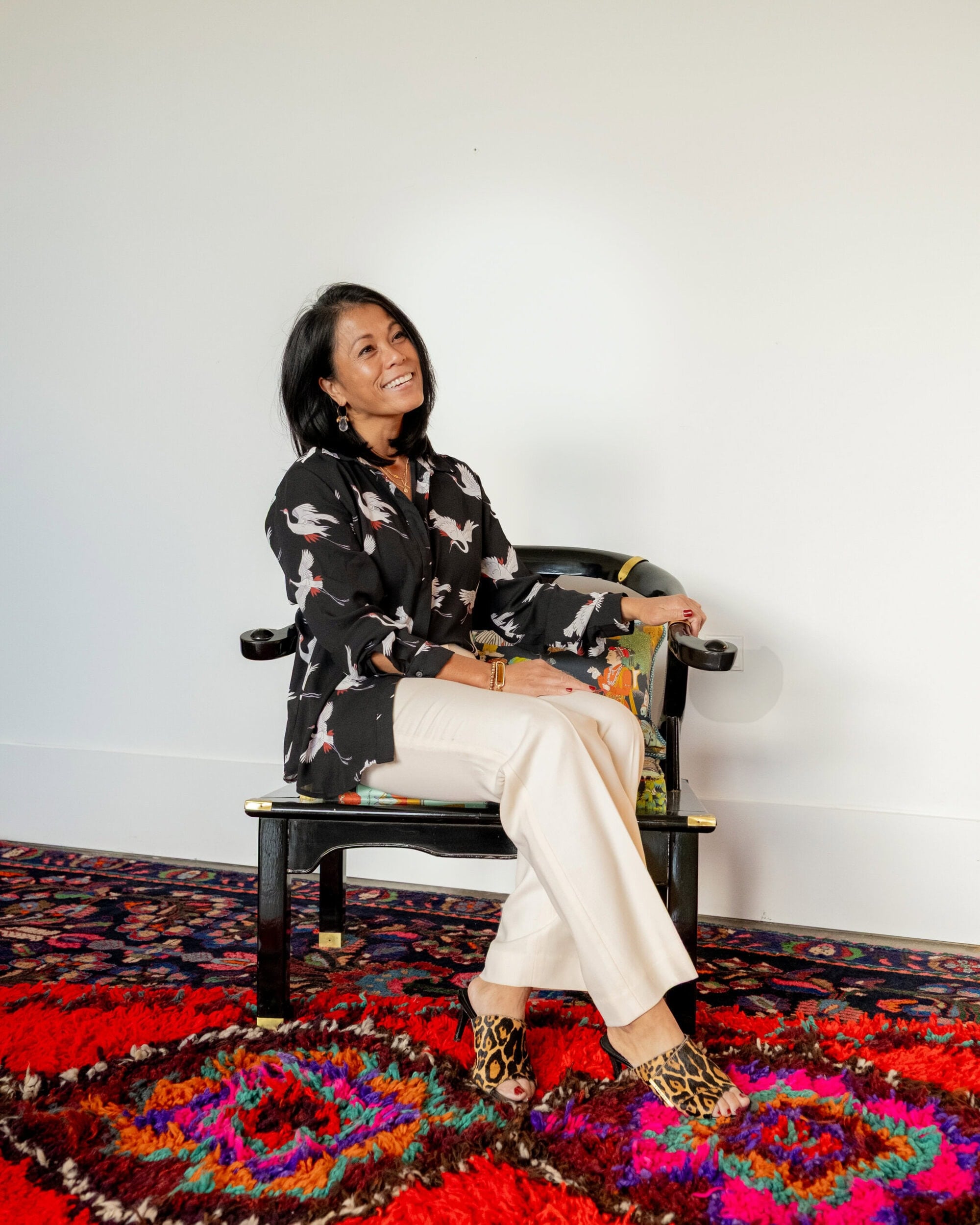 A woman sits on a chair against a plain wall, wearing a patterned black top, light pants, and leopard print shoes, on a colorful patterned rug.
