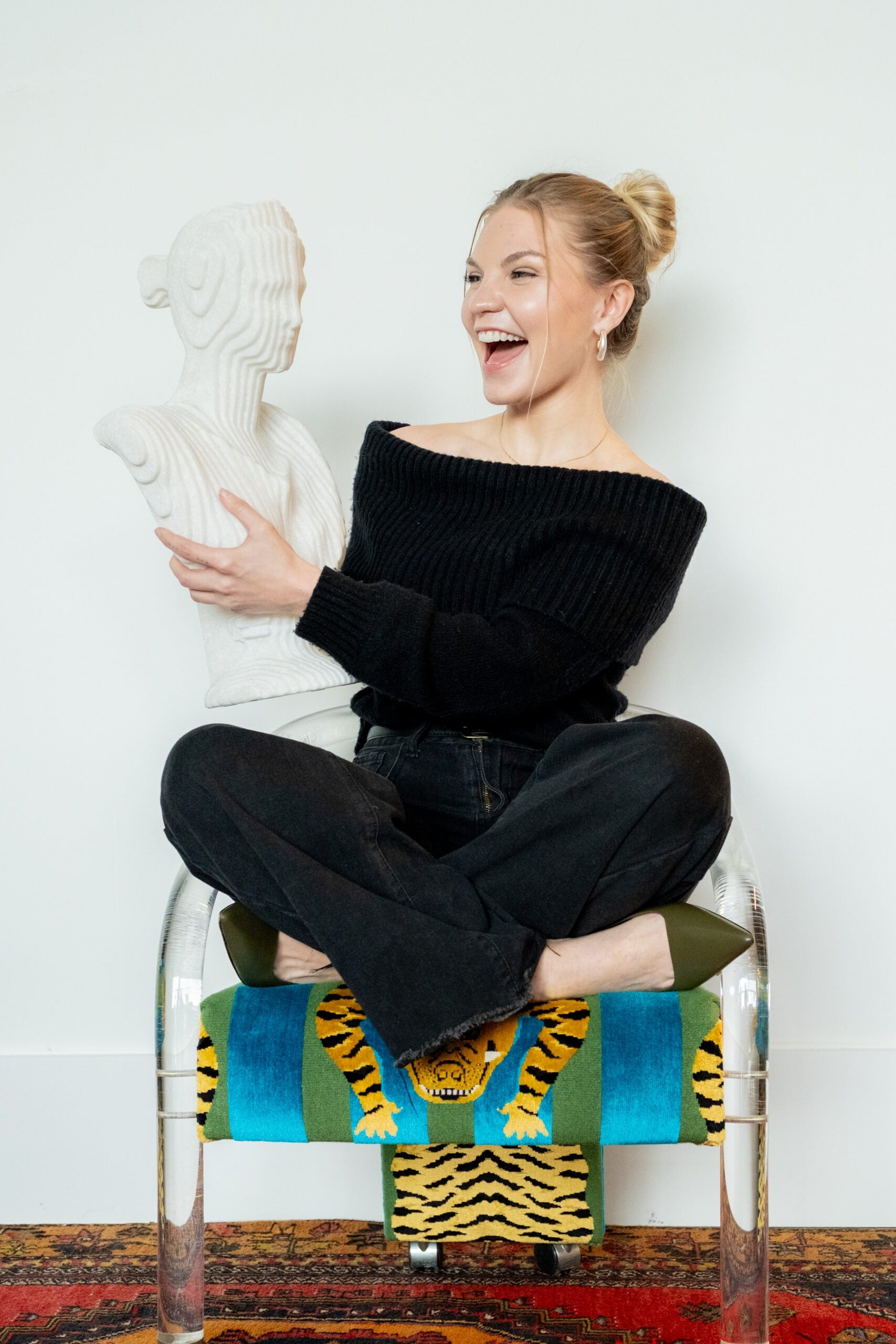 A woman sits cross-legged on a chair, smiling and holding a white bust. The chair has a colorful tiger-patterned blanket draped over it.