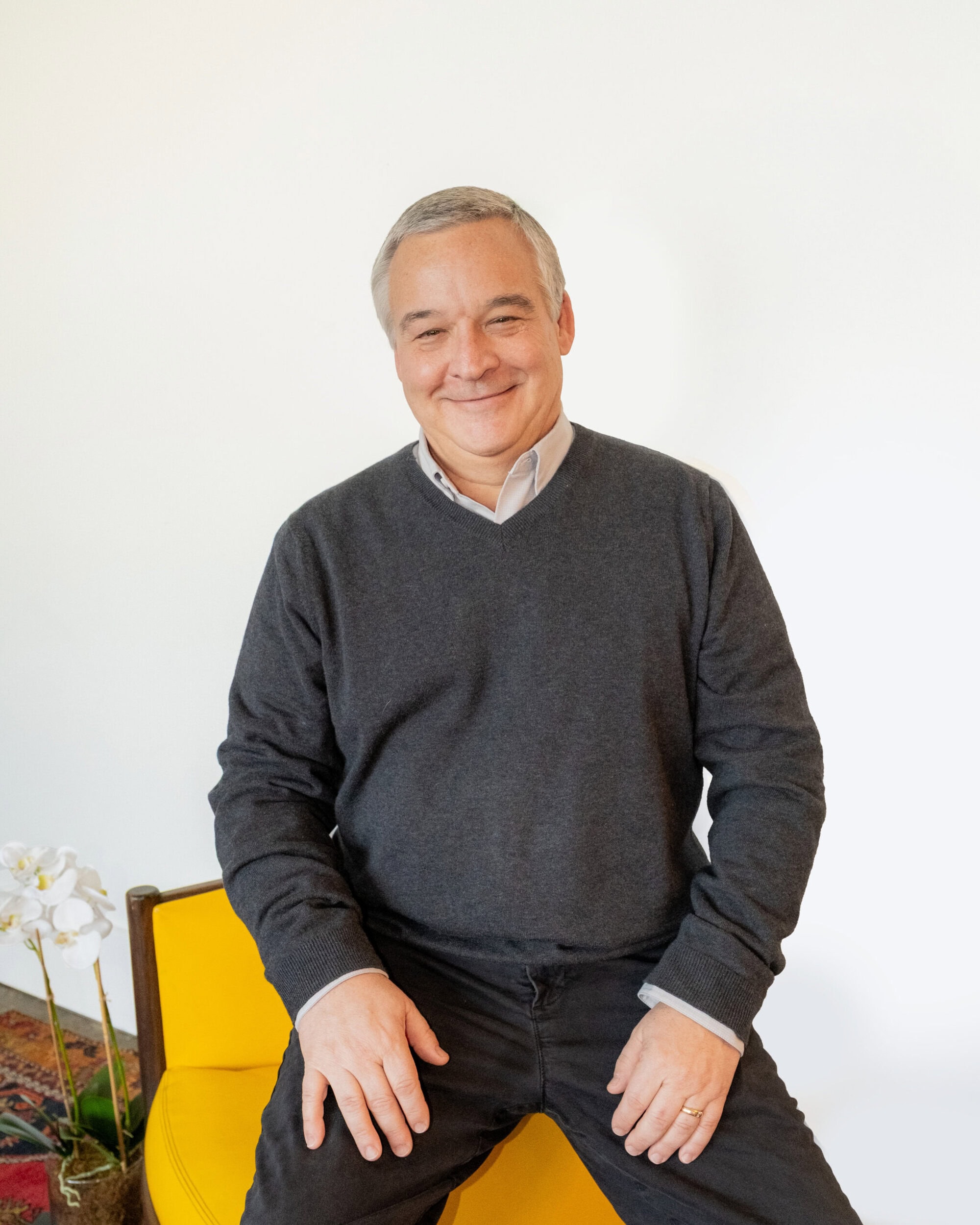 A middle-aged man with gray hair sits on a yellow chair, wearing a gray sweater and black pants, smiling at the camera. A white orchid is visible in the corner.
