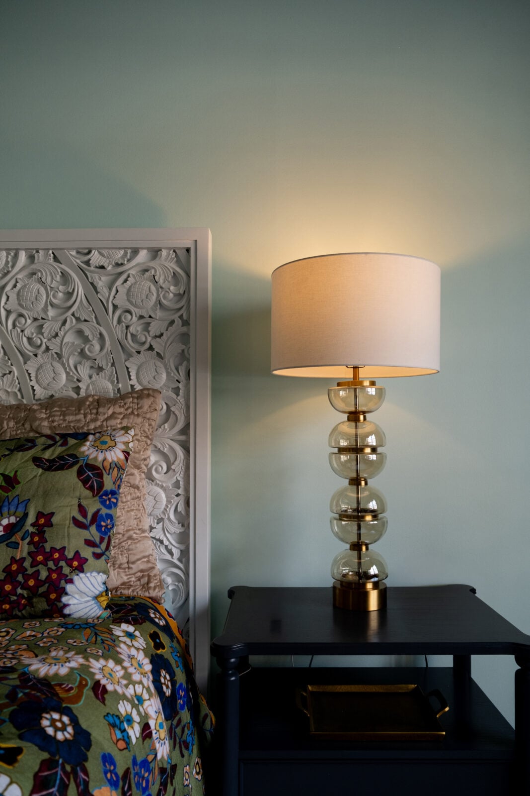 A decorative bed, a floral-patterned pillow, and a gold glass lamp with a white shade are next to a black nightstand against a light green wall.