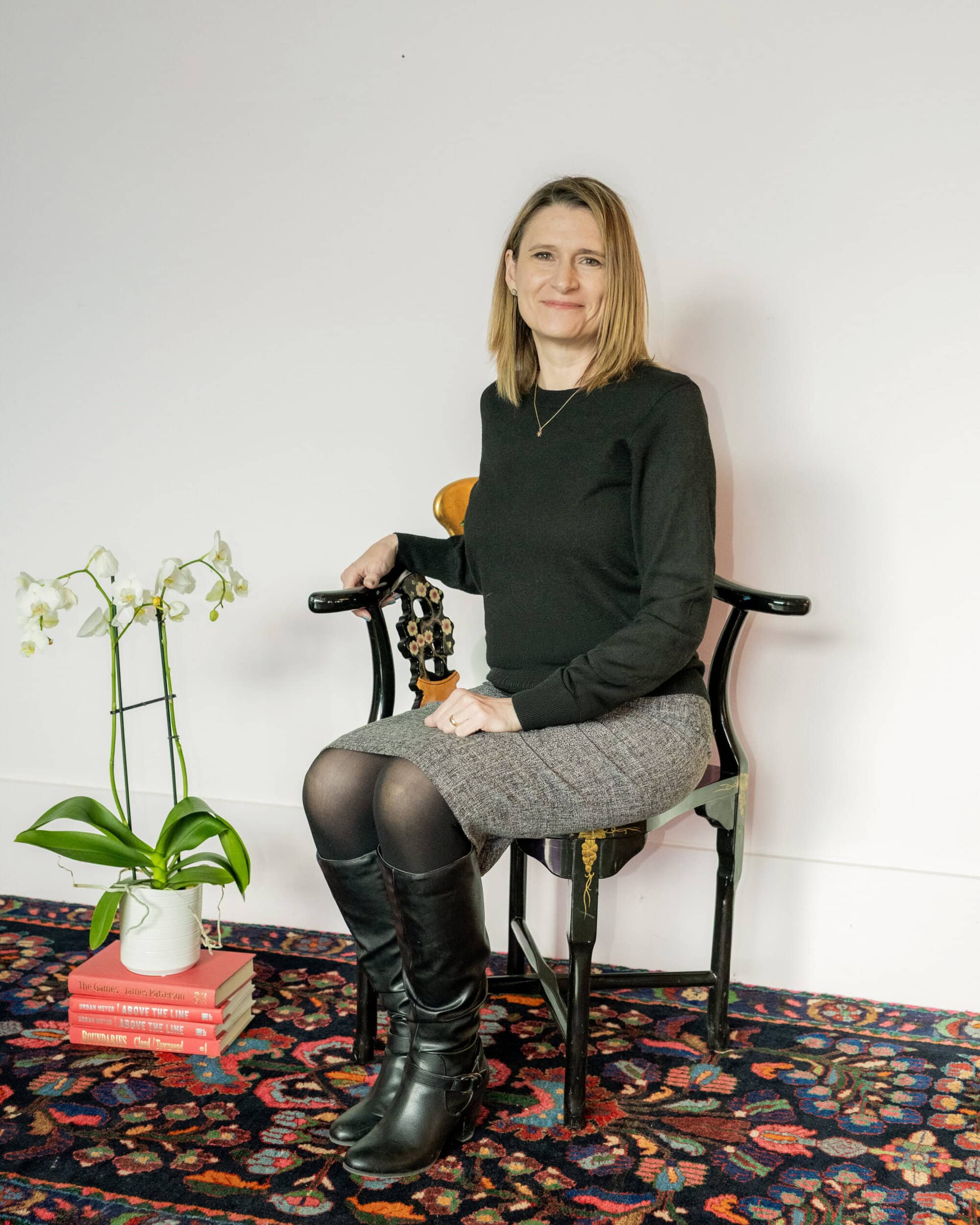 A woman sits on a black chair against a white wall, next to a potted orchid placed on a stack of red books on a patterned rug.