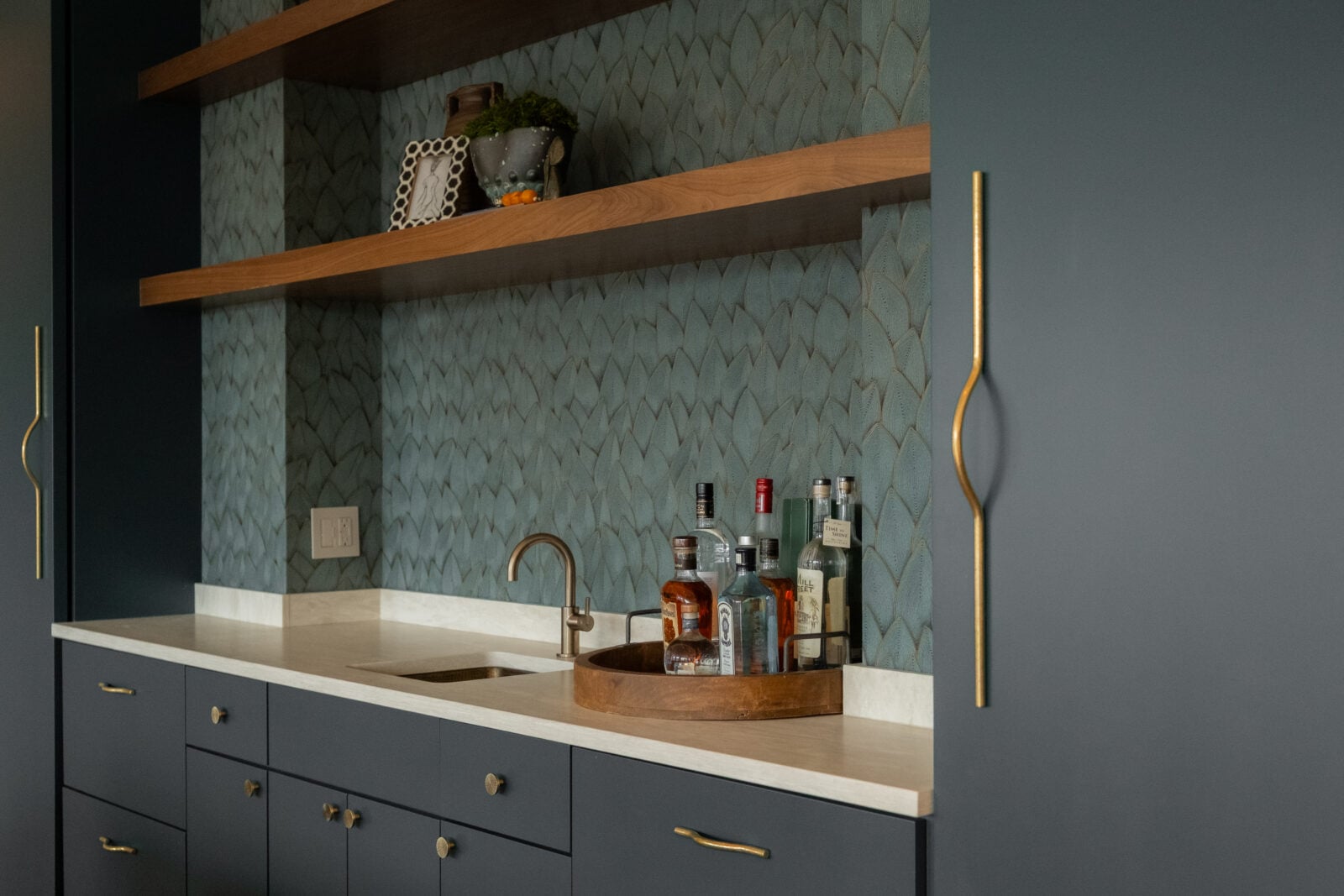 A home bar area with a small sink, bottles of liquor on a wooden tray, dark cabinets, brass handles, and open wooden shelves against a patterned backsplash.