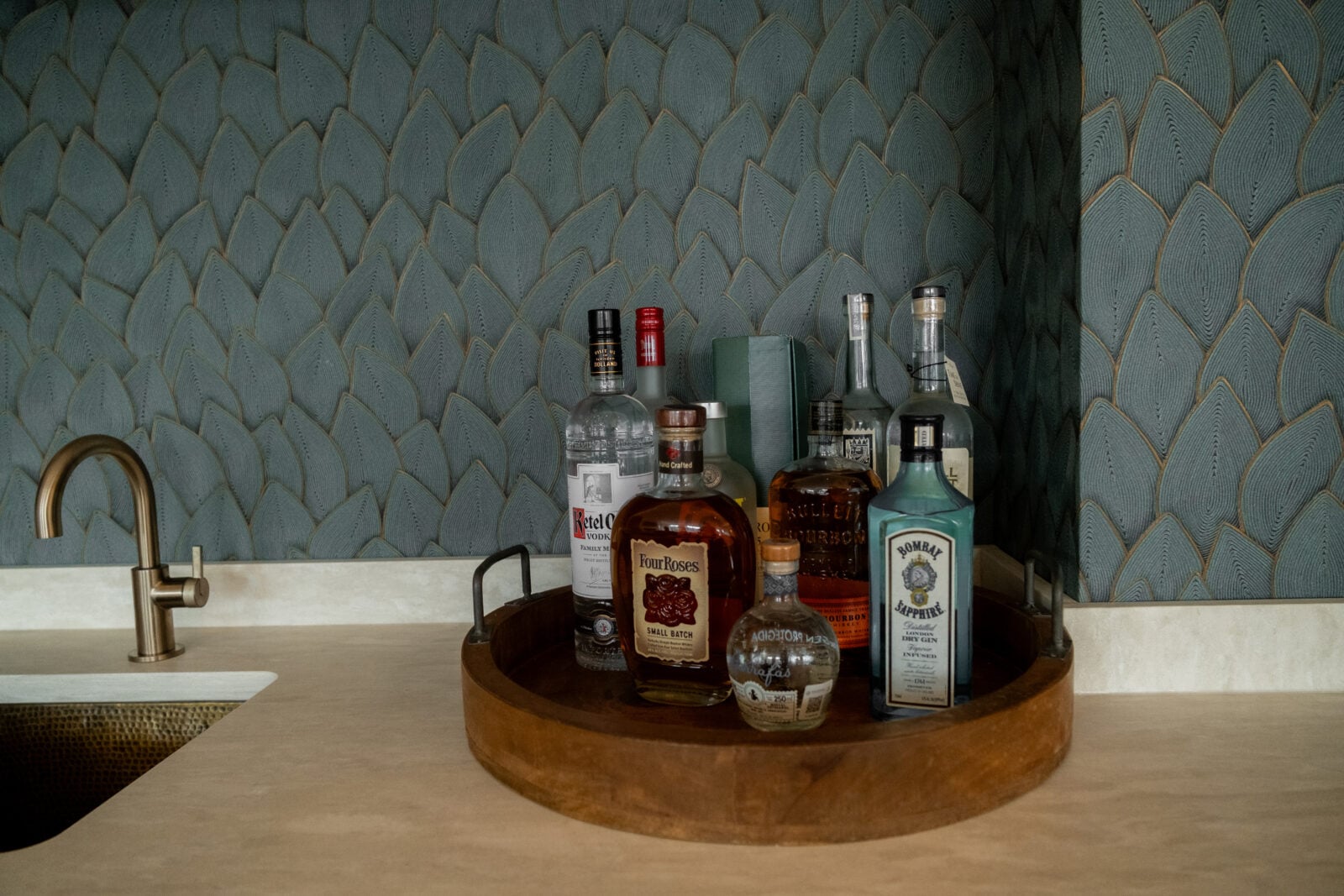 A round wooden tray holds various bottles of liquor on a beige countertop next to a brass sink, set against a blue patterned wallpaper.