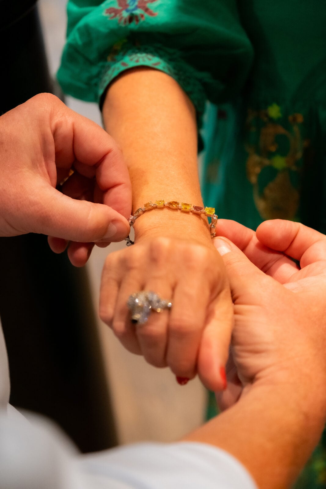 A person is fastening a gold bracelet with colored stones onto another person's wrist, who is wearing a green sleeve and a large ring.