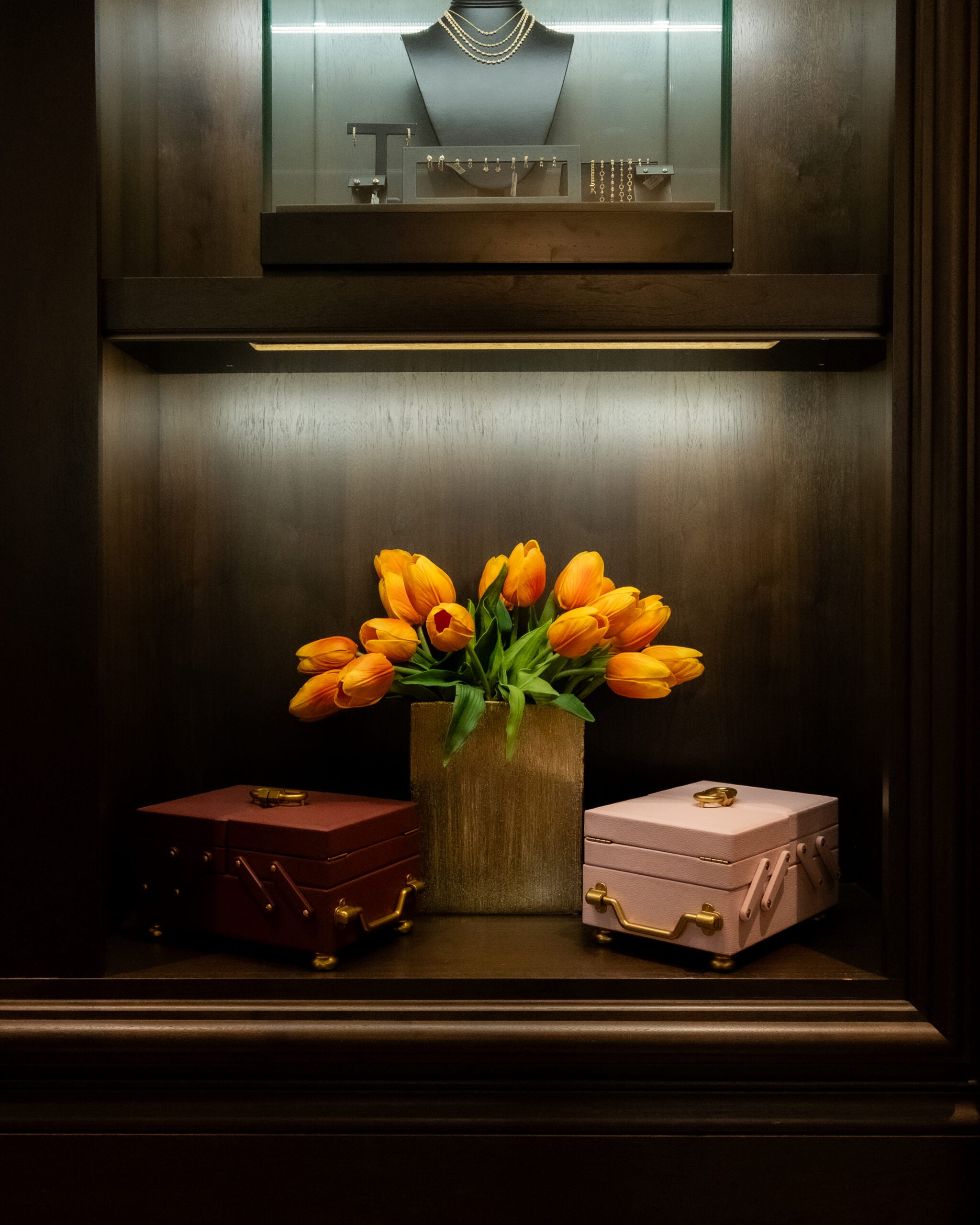 A vase of orange tulips is placed between a closed maroon box and a closed pink box, with jewelry displayed on a shelf above.