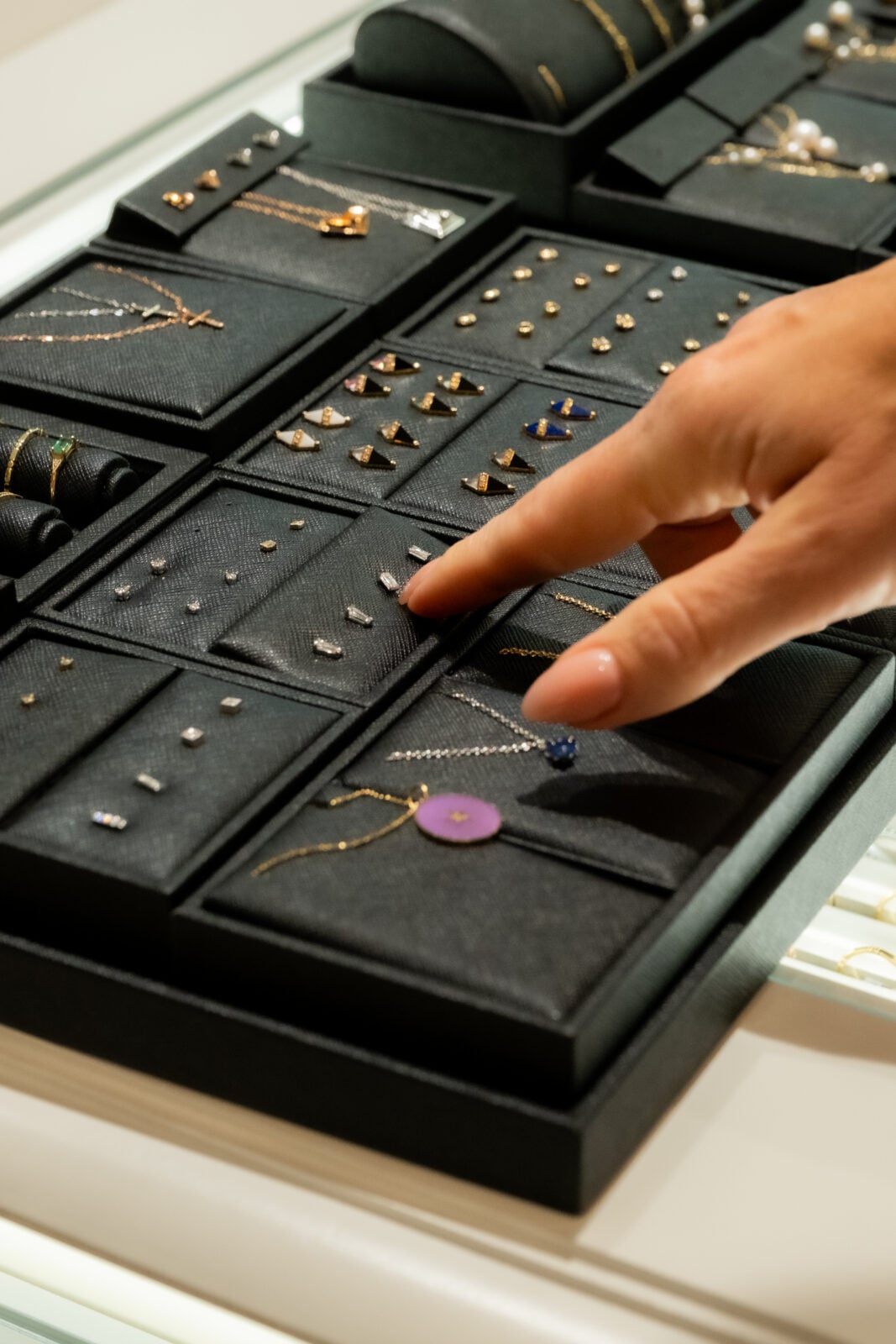 A hand points at jewelry pieces displayed in a black tray, including rings, necklaces, and earrings, inside a glass showcase.