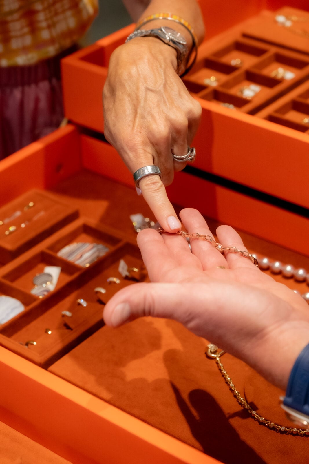 A person points to a gold bracelet in another person's hand, with various jewelry pieces displayed in an orange jewelry tray.