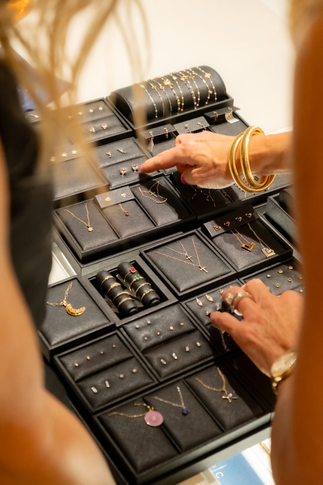 Two people view and point at a display case filled with assorted gold jewelry, including rings, necklaces, and earrings, organized in black trays.