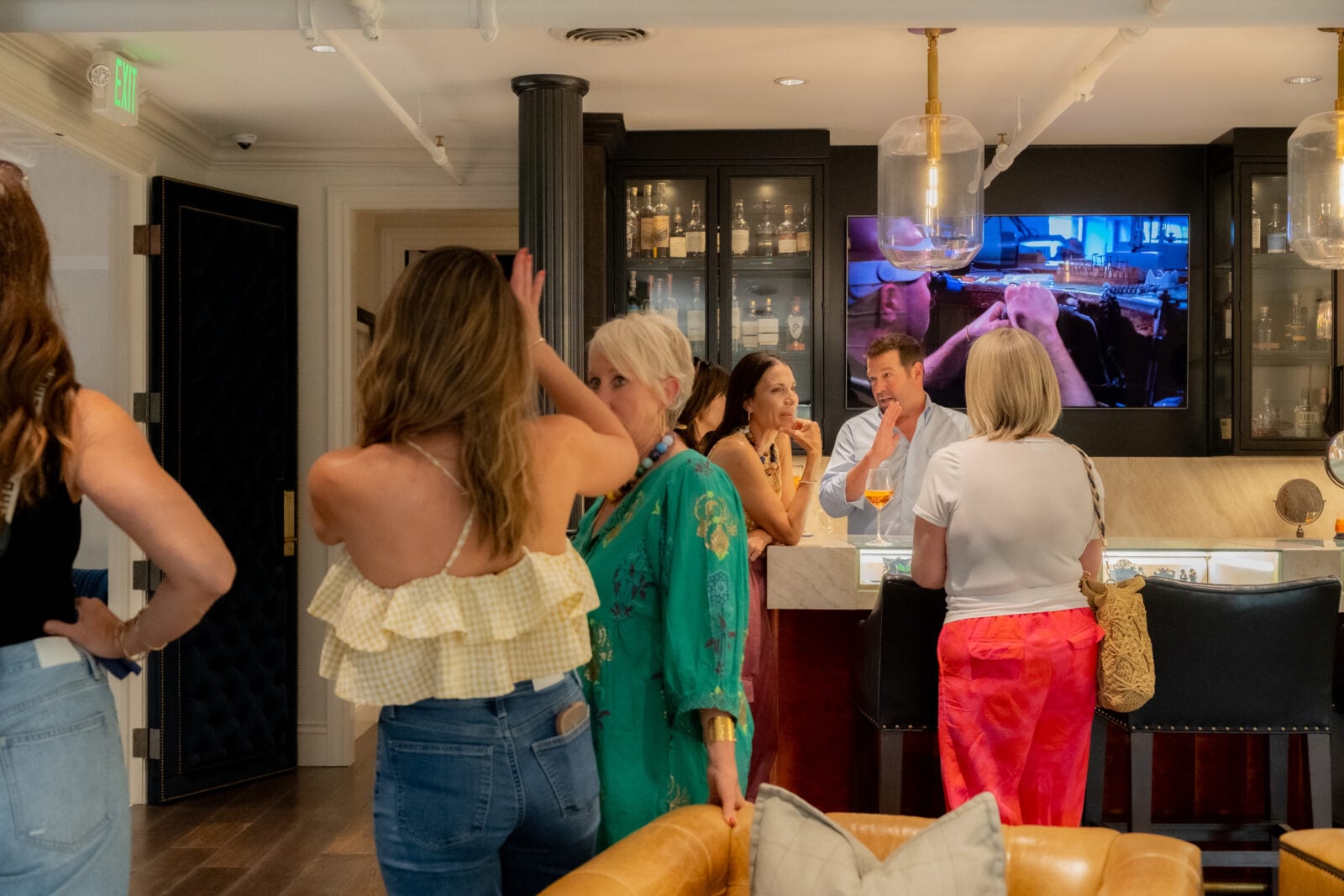Several people stand and talk near a bar counter indoors; shelves with bottles and a TV showing a rodeo scene are visible in the background.