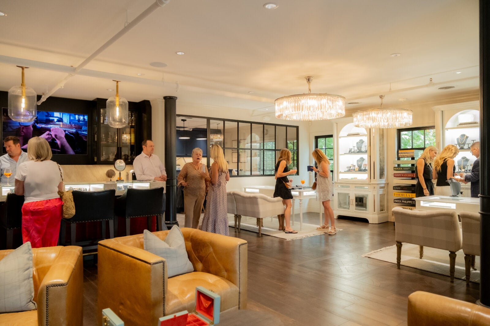 People socialize in a well-lit, upscale showroom with jewelry displays, leather chairs, and chandeliers.