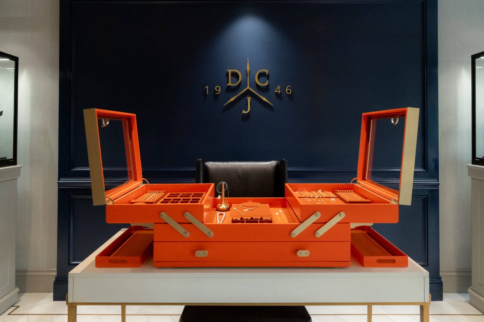 Orange jewelry box with open compartments sits on a table in front of a dark blue wall with gold "DCJ 1946" logo and a black chair.