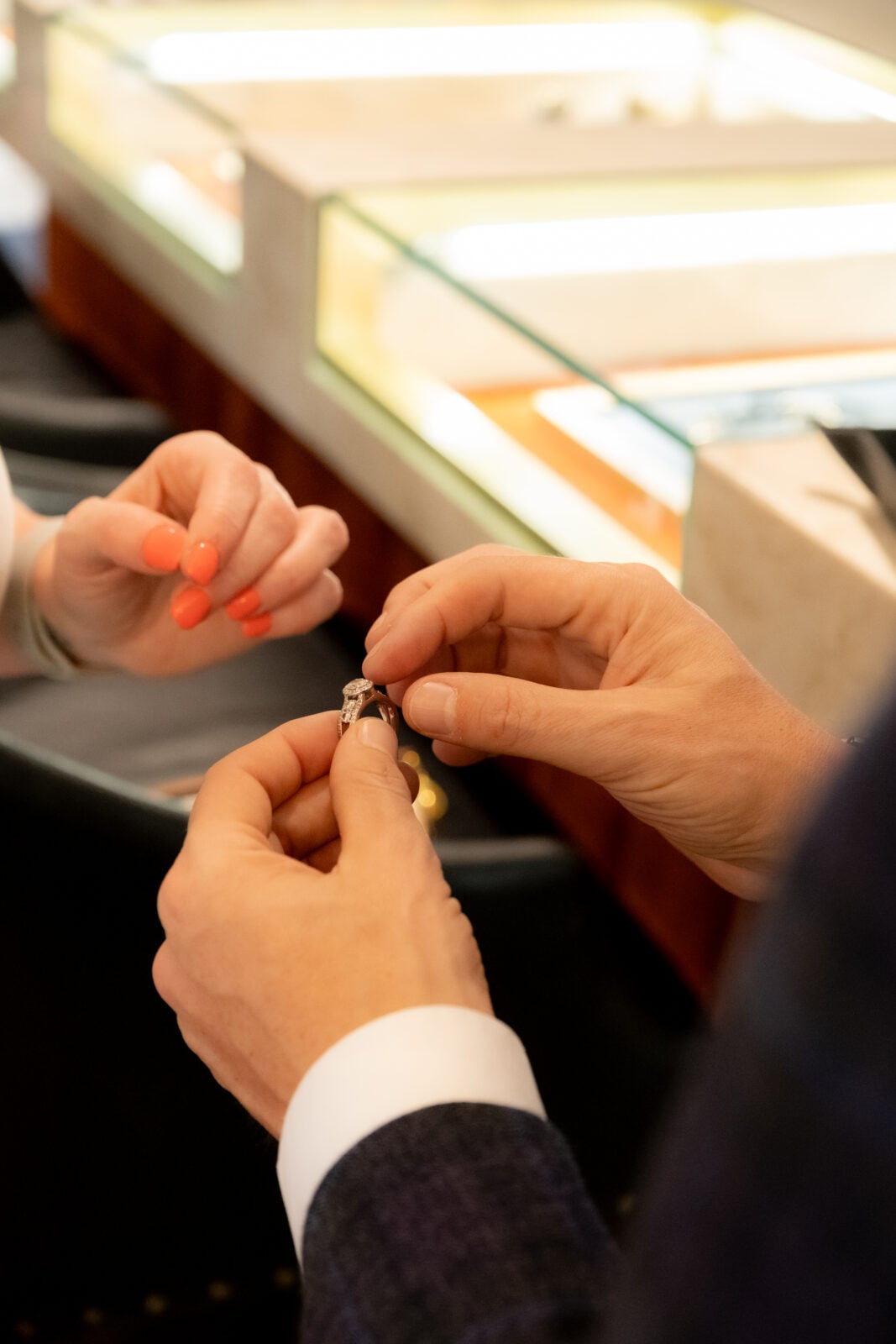 Two people exchange a ring across a jewelry store counter, with display cases visible in the background.