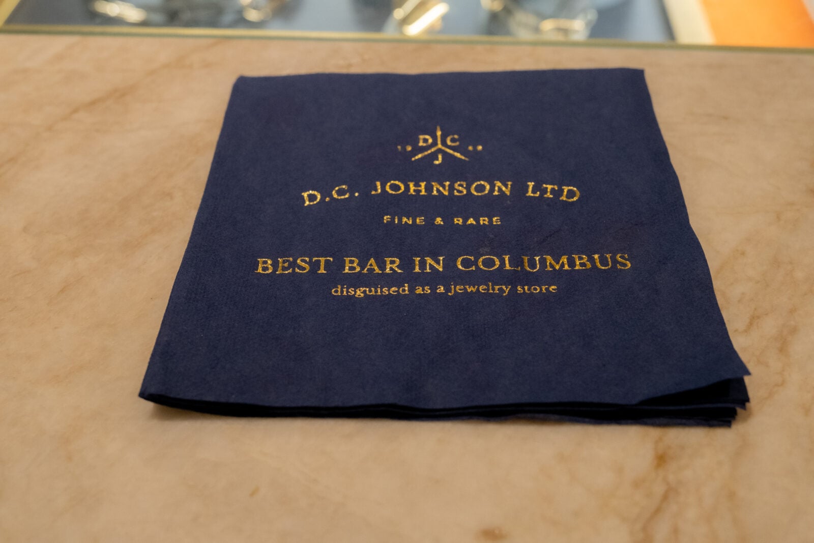 A navy blue napkin on a marble surface with gold text reading "D.C. Johnson Ltd - Best Bar in Columbus disguised as a jewelry store.