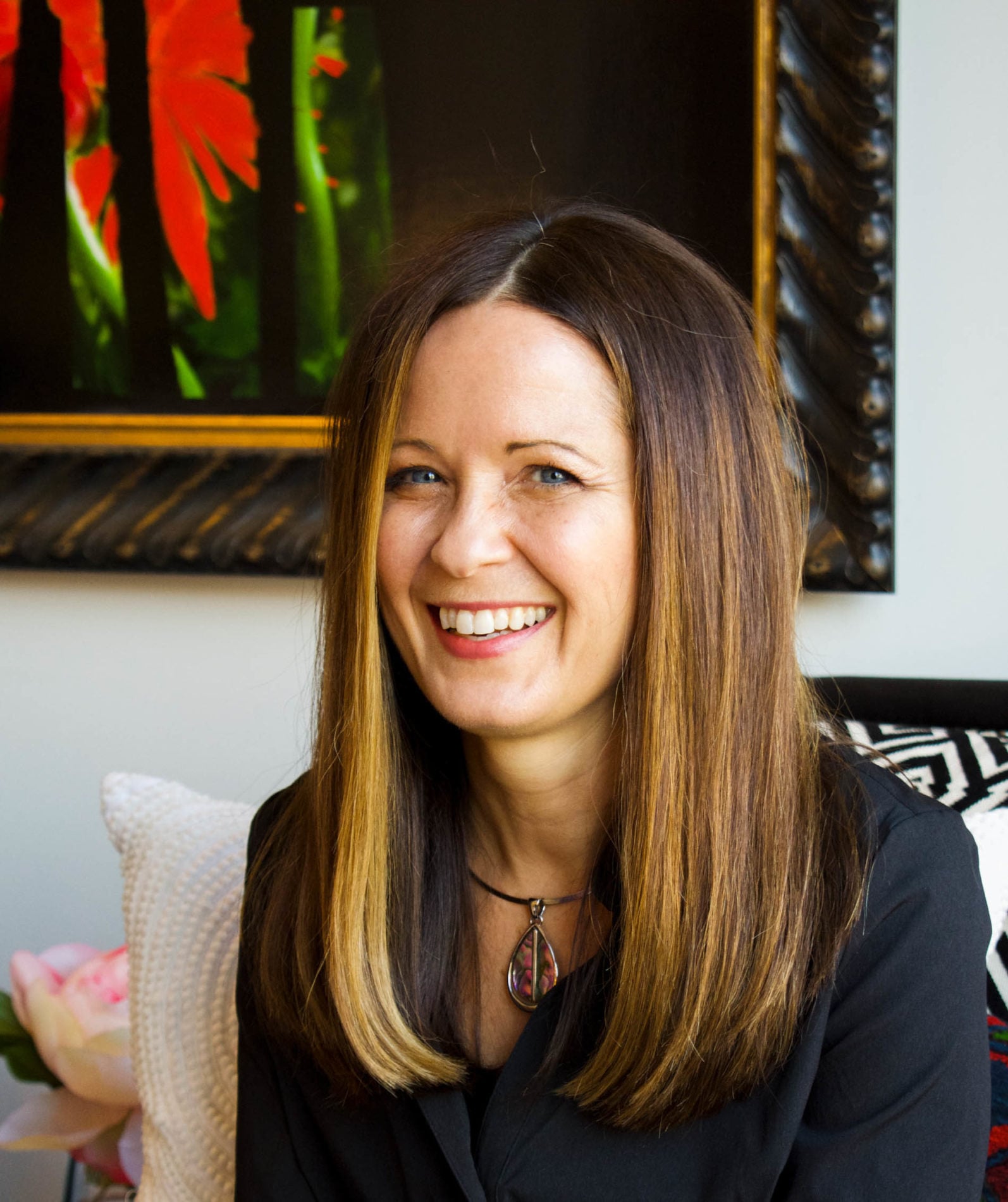 A woman with straight, long brown hair and a necklace smiles while sitting indoors in front of a framed, colorful painting—a vibrant interior reminiscent of Crimson Design Group’s signature style.