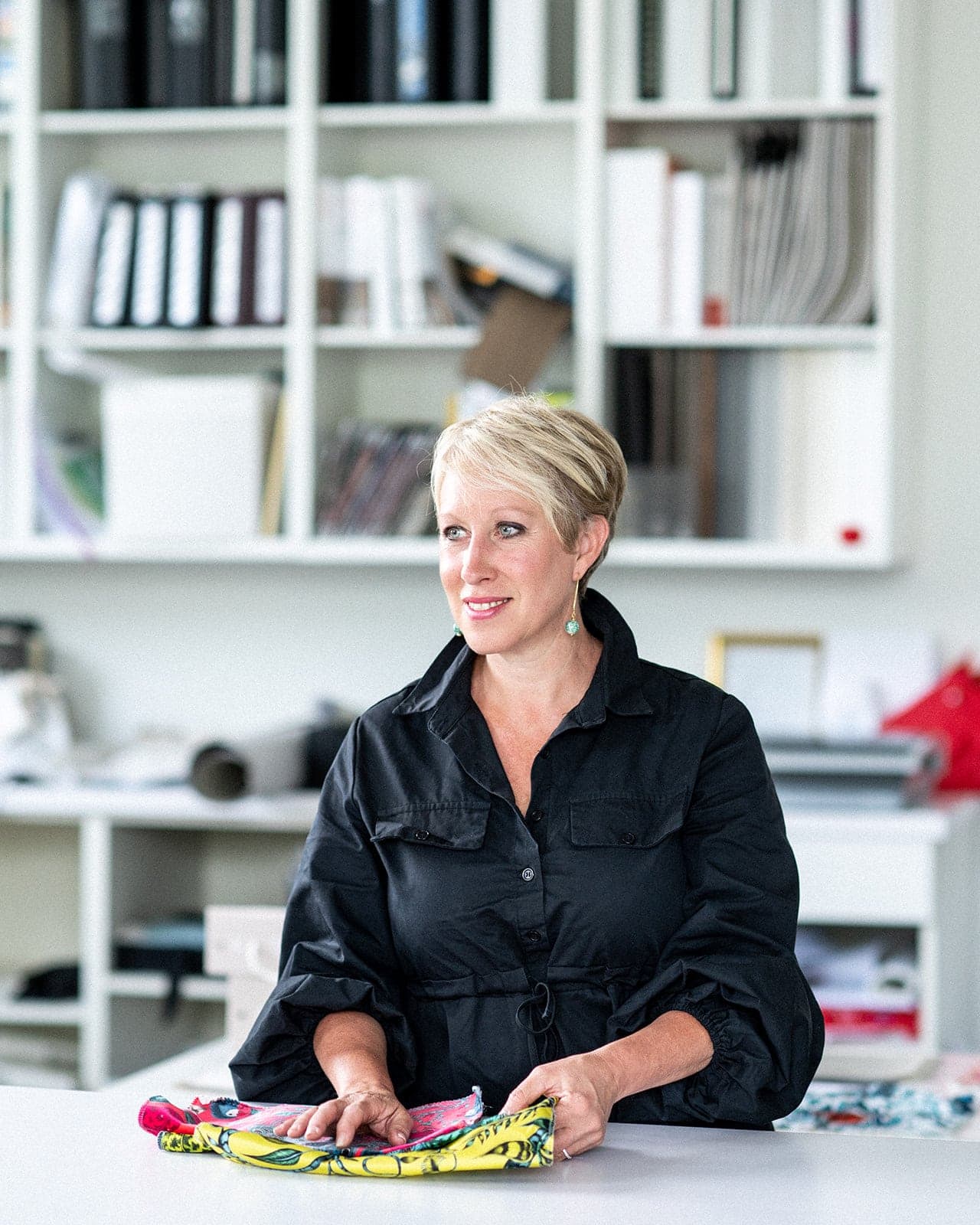 A woman with short blonde hair sits at a desk holding colorful fabric, with shelves of books and files in the background, evoking the creative spirit of Crimson Design Group.