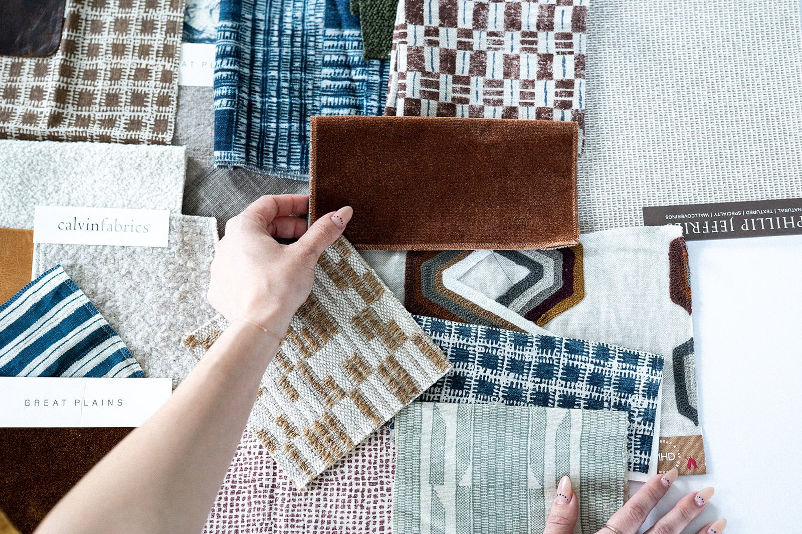 A person arranges various fabric swatches with different patterns and textures, some labeled with Crimson Design Group brand tags, on a flat surface.