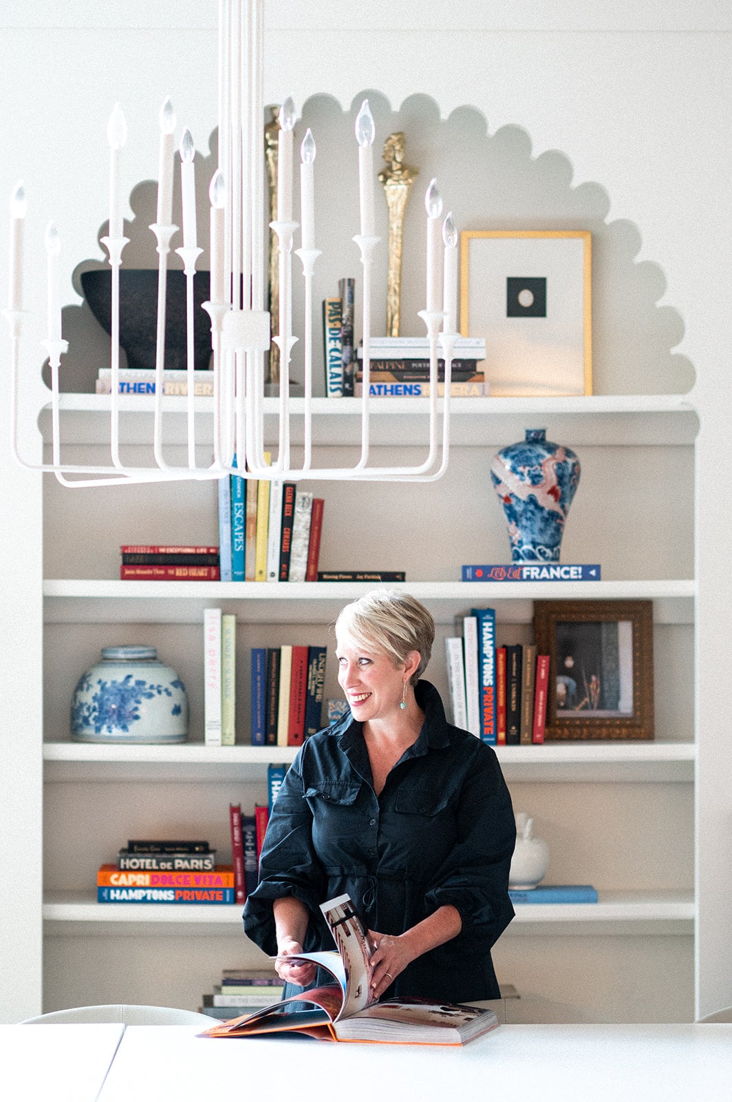 A woman with short blonde hair stands at a table, flipping through a book. Behind her, bookshelves and decorative vases showcase the signature elegance of Crimson Design Group beneath a striking white chandelier.