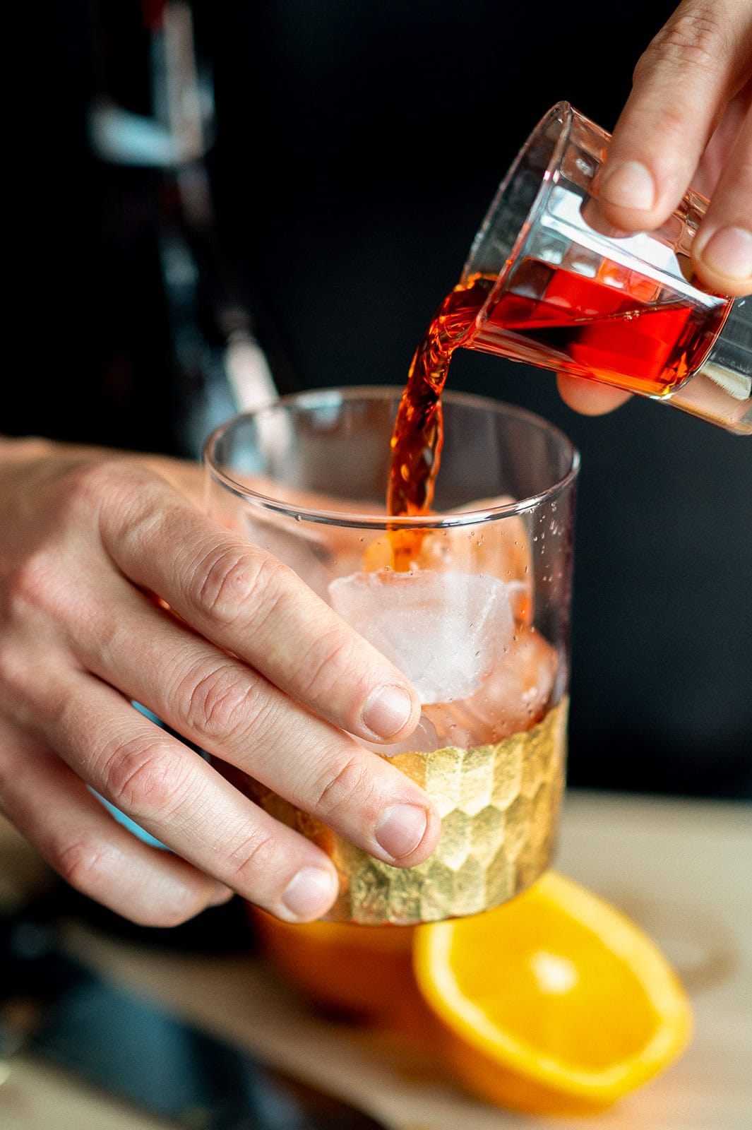 A person pours a red liquid from a small glass into a gold-patterned tumbler with ice, next to a halved orange.