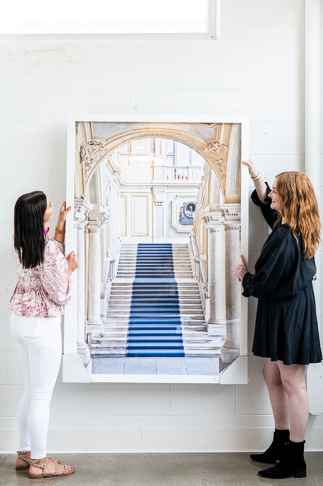 Two women stand on either side of a large framed photograph of a grand staircase with a blue carpet, mounted on a white wall—an elegant display that reflects the signature style of Crimson Design Group.