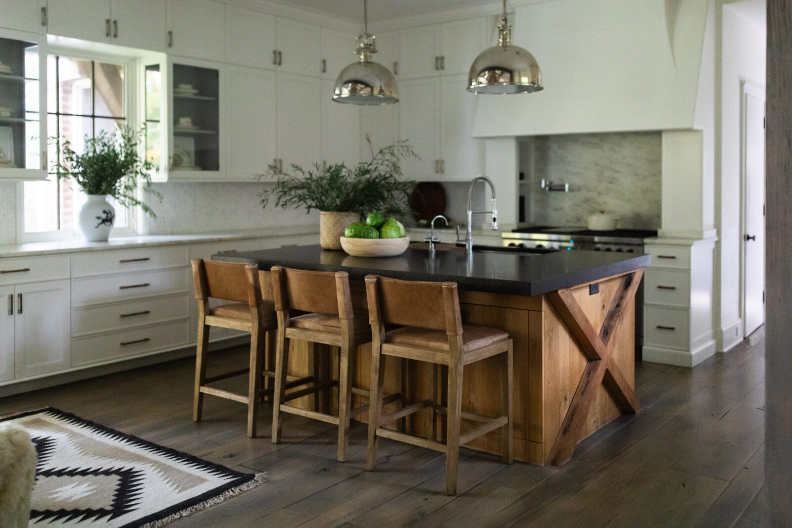 Modern kitchen with white cabinets, a black island with wooden base, three wooden chairs, pendant lights, and green plants as decor.