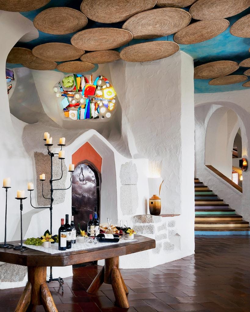 A rustic interior by Crimson Design Group with white textured walls, a wooden table set with wine and snacks, woven ceiling decor, a colorful stained glass window, and a staircase to the right.